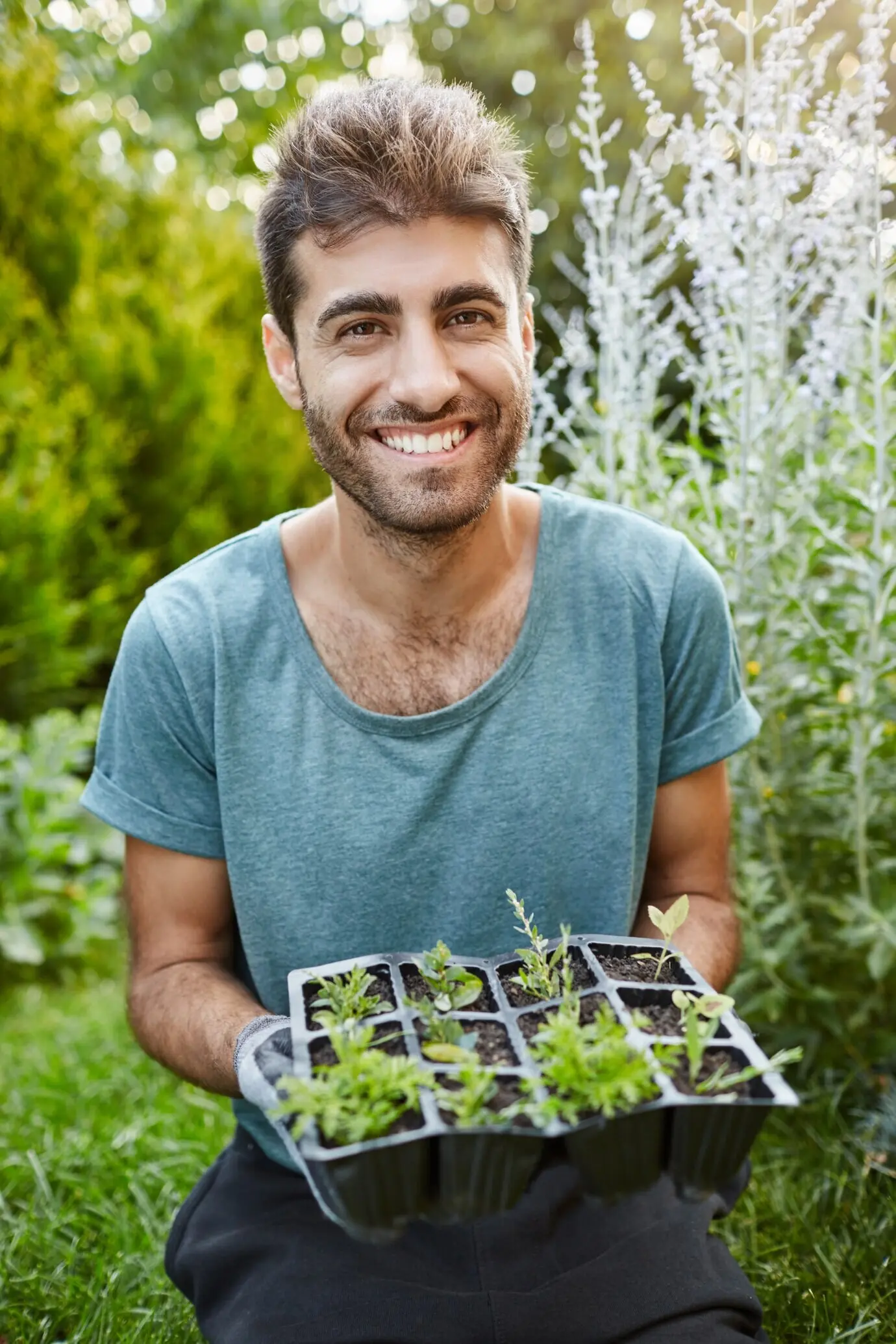 Крупный вертикальный портрет зрелого привлекательного бородатого садовника в синей футболке, улыбающегося в камеру и держащего в руках горшок с посаженными ростками.
